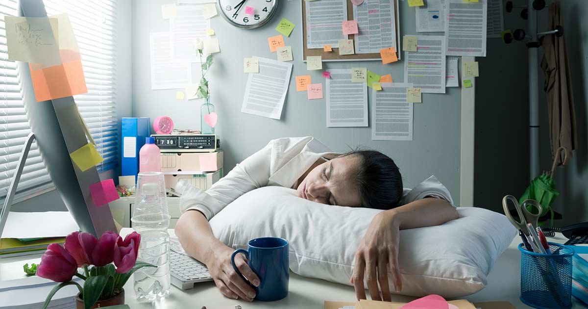 person laying over their desk sleeping while holding coffee and lots of notes in the background